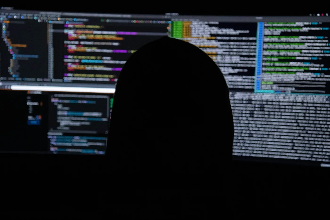 A person in silhouette in front of several monitors filled with lines of code in a dark room