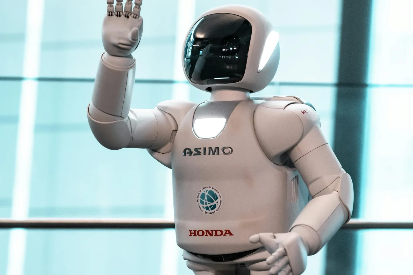Honda's white ASIMO humanoid robot raising one hand in a bright glass-walled building