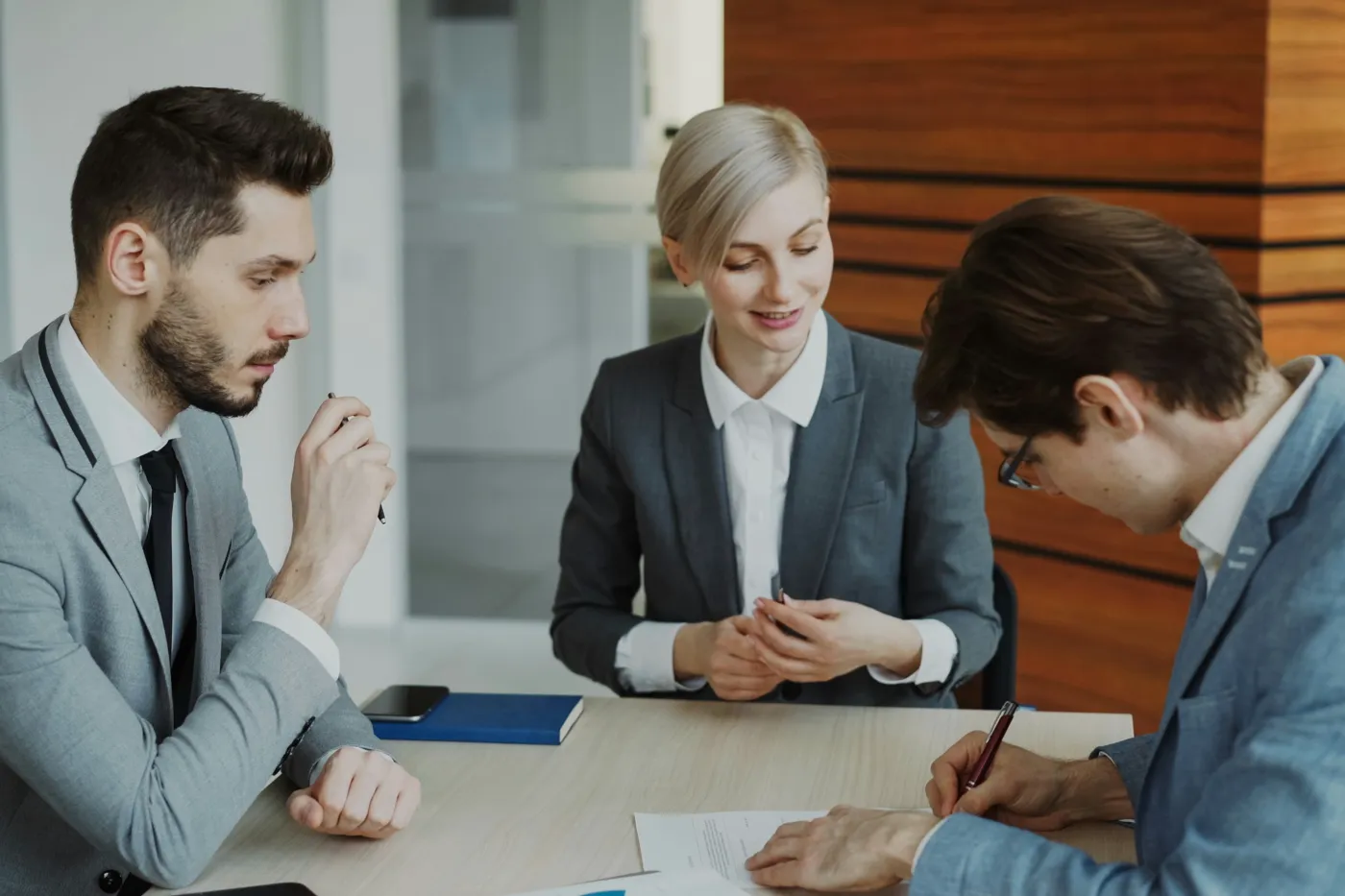 Three people at an office table, one signing a printed agreement while the others look on